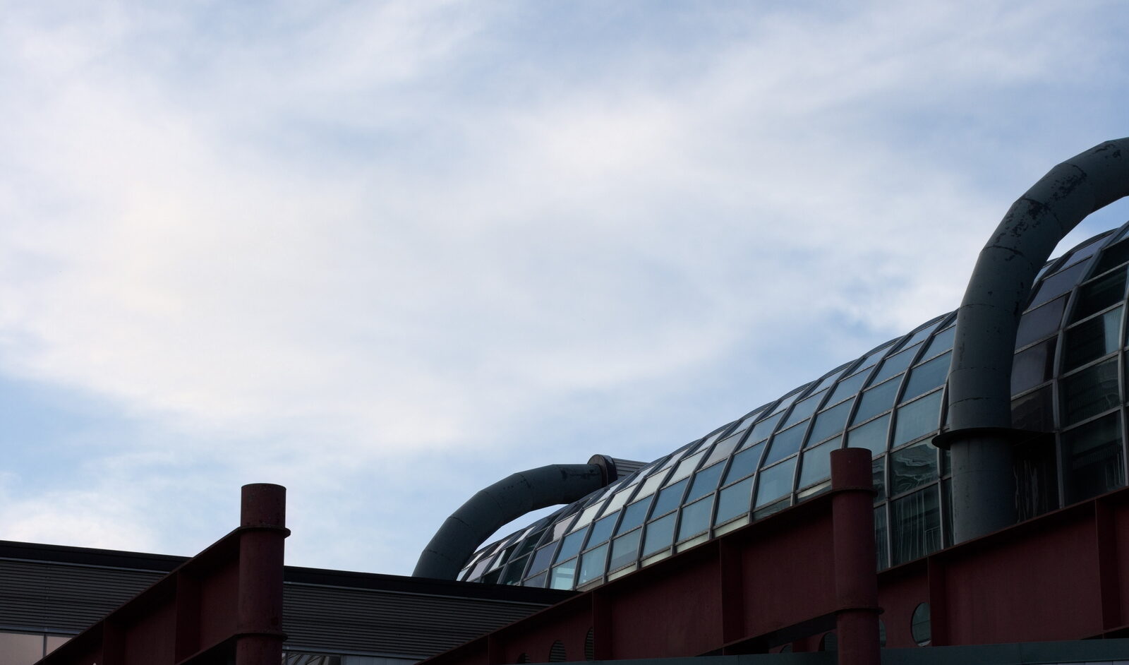 Glass-roofed building with large curved pipes against a pale sky