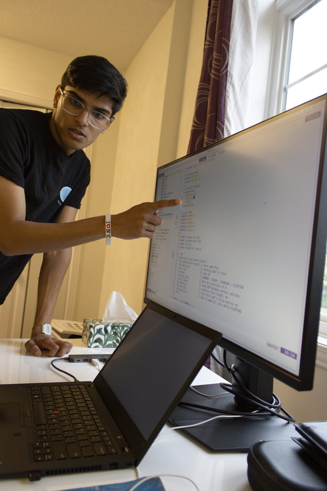Person leaning over a desk, pointing at code on a monitor next to a laptop